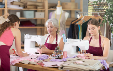 Dressmakers sew clothes next to a young fashion designer who draws a sketch of clothes on paper....