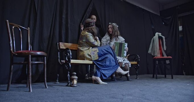 Young actors rehearse a play on a theater stage. Two actresses sit on a bench while an actor gives directions.