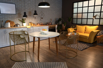 Interior of modern kitchen with glowing lamps, dining table, yellow sofa and white counters at evening