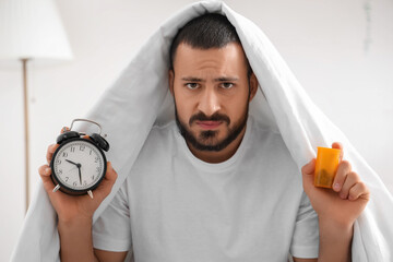Stressed young man with alarm clock and pills in bedroom