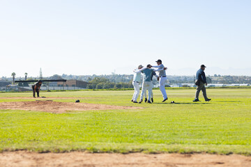 Diverse male baseball teammates celebrating win by hugging on mound at park in uniforms
