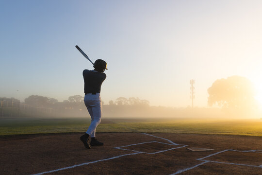Male baseball player in uniform swinging bat at home plate on infield at dawn, copy space