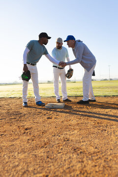 Diverse baseball coach and players fist bumping at first base with batting helmet at sunrise