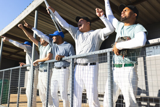 Diverse male baseball teammates cheering behind fence railing in dugout wearing caps, holding glove