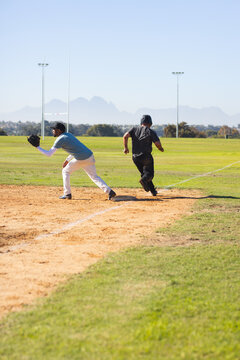 Diverse male baseball players in uniforms sprinting toward first base bag at community park diamond