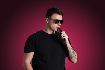 Young man in sunglasses smoking electronic cigarette on dark pink background