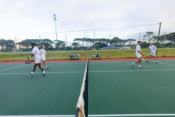 Diverse male tennis players walking across hard court at sports facility holding rackets and ball