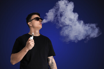 Young man in sunglasses smoking electronic cigarette on dark blue background