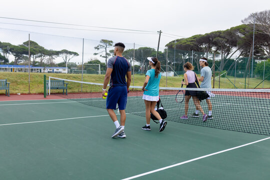 Diverse friends walking across tennis net on outdoor tennis court carrying rackets balls and bag
