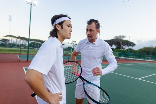 Diverse male coach and player discussing technique on tennis court with rackets and ball