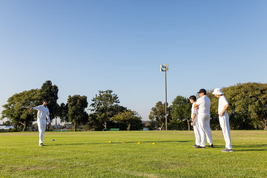 Diverse male teammates practicing cricket bowling on field with red ball and cones, copy space