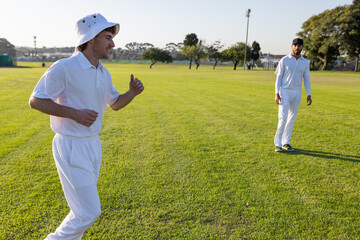 Diverse male teammates wearing cricket uniforms with bucket hat jogging on grass field, copy space