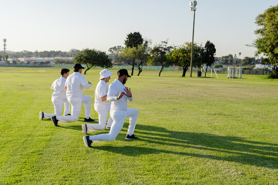 Diverse male cricket teammates performing forward lunges on cricket field wearing uniforms and caps - Powered by Adobe