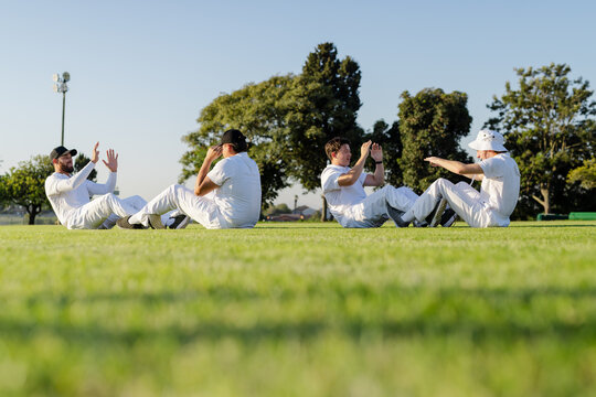 Diverse male teammates doing sit-ups and high-fiving on grass in cricket uniforms under floodlight - Powered by Adobe