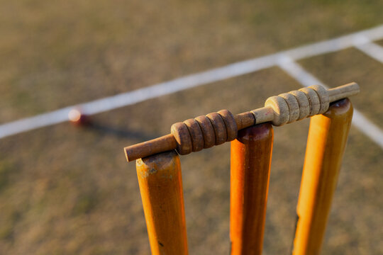 Three wooden cricket stumps are standing on cricket pitch with bails, red ball near crease lines