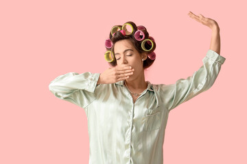 Sleepy young woman in pajamas with hair curlers on pink background