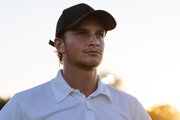 Man gazing across golf course at sunset, wearing black baseball cap and white polo shirt