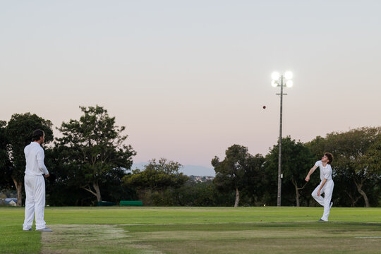 Diverse male teammates bowling red cricket ball and fielding on pitch under floodlight, copy space - Powered by Adobe