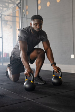 African American man kneeling on mat in gym with watch grasping two yellow accent kettlebells