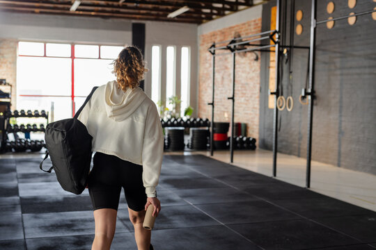 Female gym-goer walking through gym in black cycling shorts holding foam roller and duffel bag