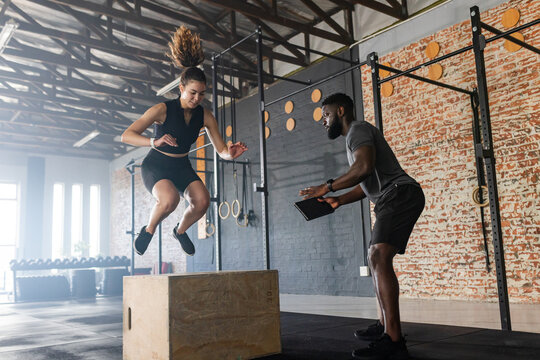 Diverse trainer and trainee performing plyometric box jump at warehouse gym with tablet, copy space