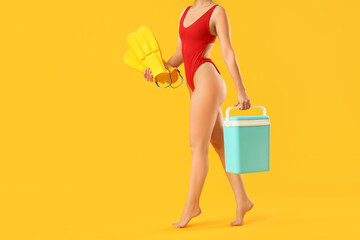 Young woman in swimsuit, with swimming flippers and portable fridge on yellow background