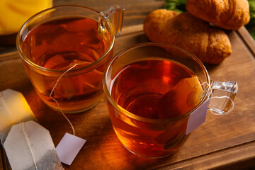 Cups of hot beverage with tea bags and croissants on wooden background