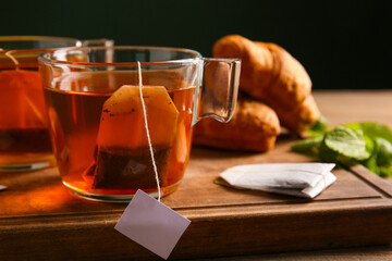 Cups of hot beverage with tea bags, mint leaves and croissants on wooden table against dark wall