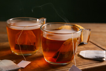 Cups of hot beverage with tea bags on wooden table against dark wall