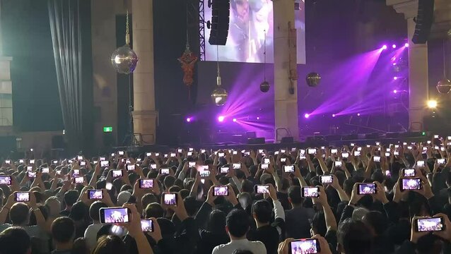 Enthusiastic crowd at a concert captures the vibrant live performance on smartphones, illuminated by dynamic purple stage lights, showcasing modern event attendance and digital engagement.