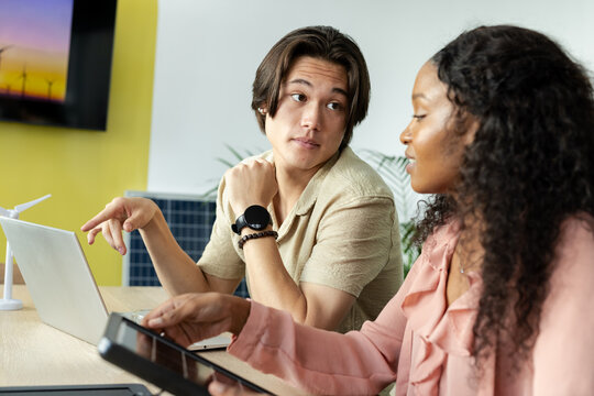Diverse coworkers discussing green energy at office desk with laptop turbine model and solar panel