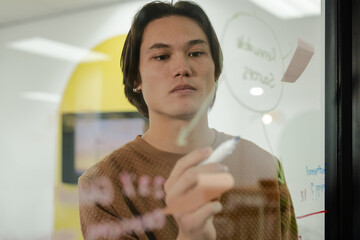 Asian man drawing circular diagram on clear glass whiteboard in office with marker and sticky note