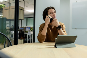 Asian man holding smartphone while thinking at round conference table with tablet folio case