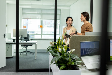 Asian coworkers holding tablet while discussing renewable energy on laptop beside planter at desk