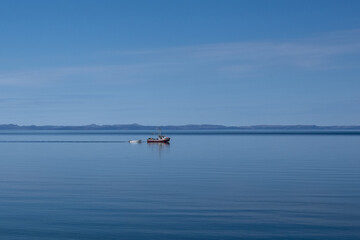 A small fishing boat glides across the calm, blue ocean, with a colorful blue sky. There are thin clouds over a mountain landmark. The red fishing boat is towing a small white open rowboat or skiff. 