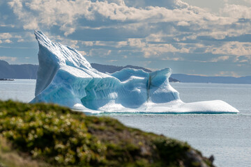 A large white iceberg formation floats in the cold ocean, its layers of textured ice and snow. The ice is in transition, melting from the warm rays of the sun at sunset. The berg is a blocky shape.