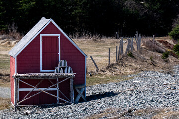 An old vintage fishing stage with a single wooden door on the top floor of the building. There's a flake for drying fish in front of the storage shed with lobster pots. There's a meadow in the back.
