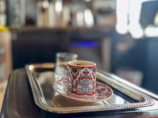 An empty Turkish coffee cup, kahve fincani, and saucer on a silver tray. The colorful porcelain cup has a thin red rim. The tray has a small glass of water next to the colorful mug. 