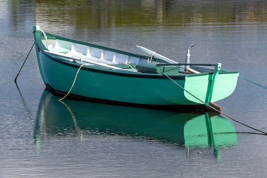 A bright green wooden boat with dark green trim, known as a Rodney boat or a go-ashore vessel. The center of the boat is white and there's two oars in it. The small boat is moored with rope in a pond.
