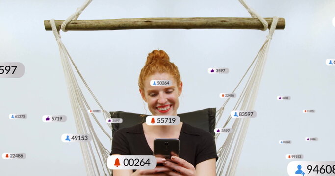 Smiling red-haired woman holding phone in rope wooden hammock chair at studio with notifications - Powered by Adobe