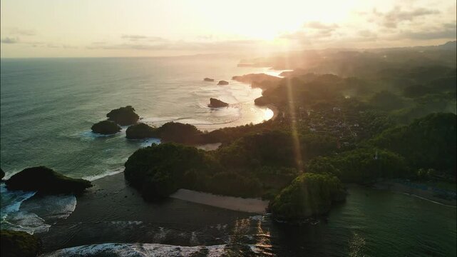 Dramatic sun rays pierce the hazy sky during a golden hour sunset over a rugged coastline dotted with forested sea stacks and hidden coves. Watu Karung Beach in Pacitan, Indonesia.