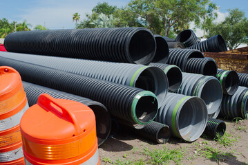 Large Drainage Pipes Stacked at a Construction Site with Traffic Barrels. Outdoor construction site featuring black and gray drainage pipes stacked, surrounded by traffic safety barrels under a sunny 