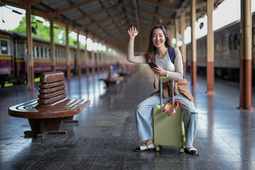 A woman is sitting on a bench with her luggage and waving at the camera