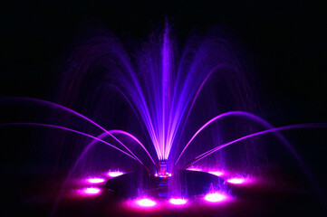 Floating fountain in neon purple blue, large detailed colorful horizontal closeup at night, lit up illuminated color changing bright city park lights, colored water jets on dark black background