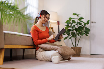 Asian Teen girl with disability wearing headphones choosing playlist for studying holding digital tablet while sitting on floor near sofa
