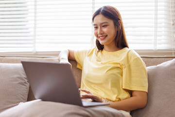 Obraz premium Concentrated asian girl sit on couch working on laptop browsing internet at home during lazy weekend, focused young woman freelancer busy using computer surfing wireless web shopping online 