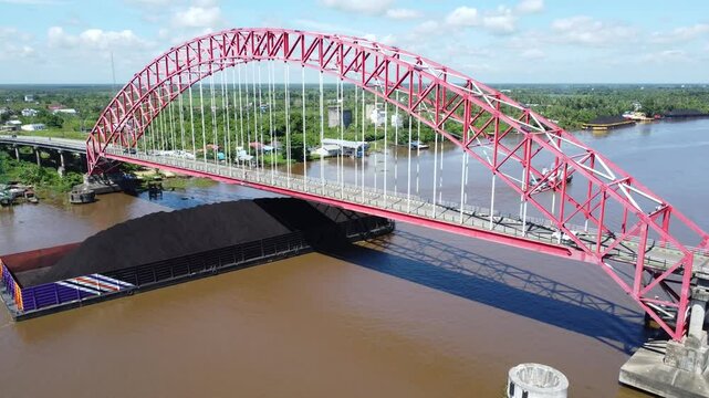 Aerial view of a tugboat pulling a barge filled with coal across the Rumpiang Bridge in the waters of South Kalimantan, Barito River