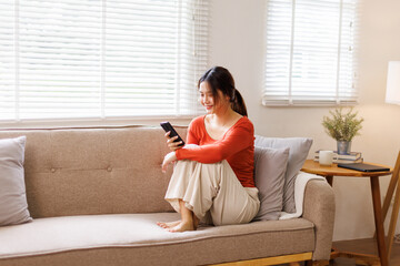 Happy young asian woman relax on comfortable couch at home texting or chatting messaging on the phone, smiling girl use cellphone, online message, shopping online from home
