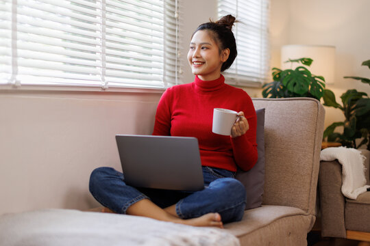 Relax Concept. Happy woman drinking coffee using pc sitting on comfortable couch at home in living room and dreaming. Cheerful casual famale resting on sofa, enjoying weekend free time.
