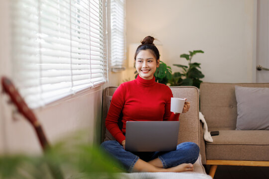 Relax Concept. Happy woman drinking coffee using pc sitting on comfortable couch at home in living room and dreaming. Cheerful casual famale resting on sofa, enjoying weekend free time.
